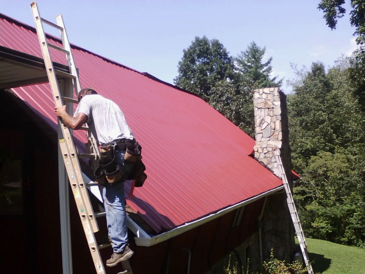Roofer on a ladder beside a red metal roof and stone chimney — Soffit & Fascia Repair by Roofing Brothers in Dover Plains
